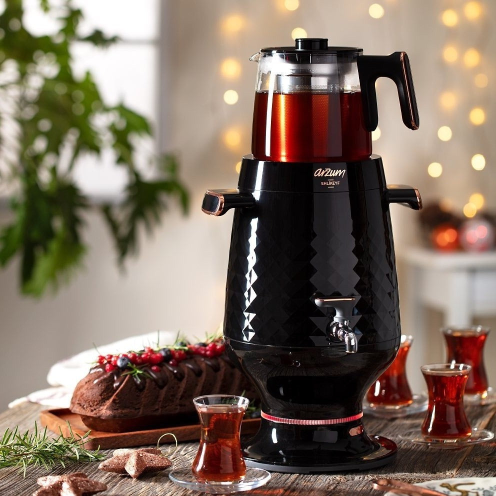 Black diamond-patterned tea maker with a glass container on a wooden table, surrounded by tea cups and a loaf of bread.